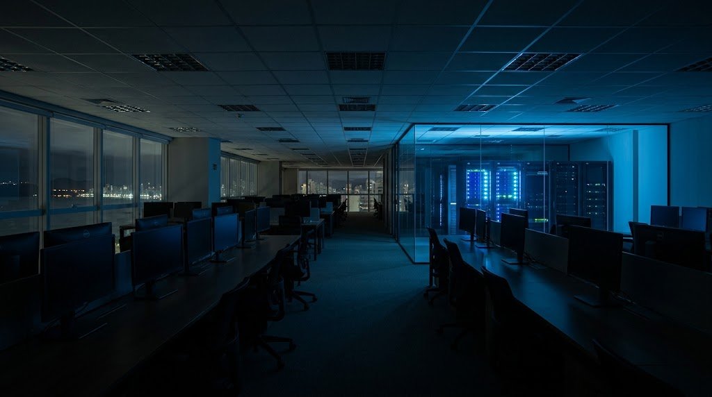 Home 7 Escritório corporativo vazio à noite com a sala de servidores acesa ao fundo, representando a janela de risco que o monitoramento 24x7 cobre.